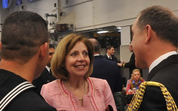 A formal military event aboard a naval vessel, likely a commissioning ceremony. A woman in a pink tweed jacket is engaged in conversation with a sailor in dress uniform bearing 'USS AMERICA' on his uniform and a senior officer with gold aiguillettes on his shoulder. The setting appears to be an interior hangar or ceremonial deck of a ship.