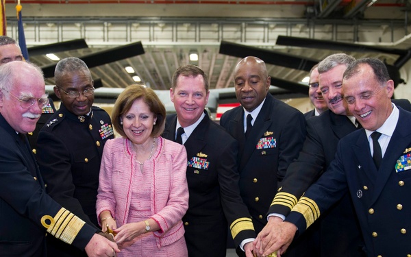 A formal military ceremony taking place in what appears to be an aircraft hangar, featuring naval officers and a civilian woman in pink cutting a large decorated cake. The cake reads 'American Virgin to Americans' with the date 'August 7, 2014,' suggesting this is a commemorative event. Multiple high-ranking naval officers wearing dress uniforms with medals and insignia are gathered around the table.