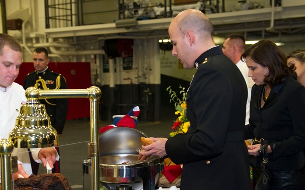 A formal military dining event or reception taking place in what appears to be a ship's hangar bay or large indoor military facility. A chef in white uniform is carving a large roast at a decorated buffet table while military personnel in dress uniforms and civilian guests in formal attire queue for food. The setting suggests an official military ball or formal mess dinner.