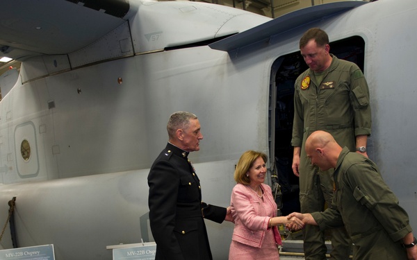 A formal military greeting ceremony is taking place inside an aircraft hangar in front of an MV-22B Osprey tiltrotor aircraft. A senior Marine officer in dress uniform and a woman in a pink suit are shaking hands with a pilot in flight suit who is descending from the aircraft. Informational display boards about the MV-22B Osprey are visible in the foreground.