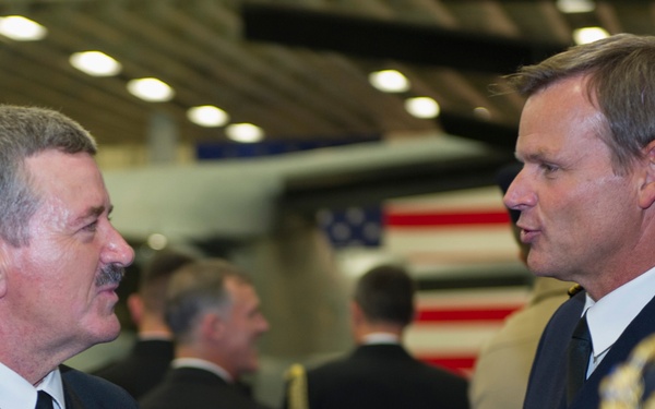 Two military officers in dress uniforms are engaged in conversation at what appears to be a formal military ceremony or reception. An American flag is visible in the background along with other attendees in formal attire. The officer on the left wears U.S. Navy dress blues with service ribbons, while the officer on the right appears to be in a foreign military dress uniform with gold aiguillettes.