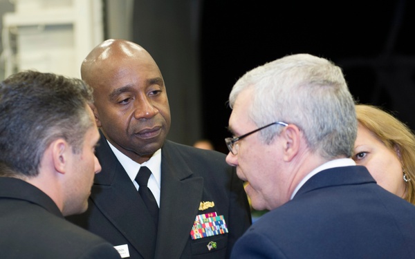A U.S. Navy officer in dress blues uniform with multiple service ribbons and gold buttons is engaged in conversation with two civilian men in dark suits at what appears to be a formal reception or military event. A woman in a black and white patterned dress is visible in the background along with other attendees. The setting appears to be an indoor venue with multiple people gathered.