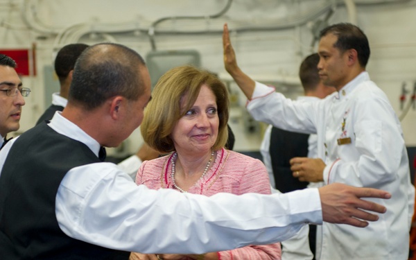 A woman in a pink blazer and pearl necklace is engaged in conversation with a man in a dark vest and white shirt in what appears to be a military vessel's hangar or interior deck. Several uniformed personnel, some in white service uniforms and others in dark dress uniforms, are visible in the background. The setting suggests an official visit or reception aboard a naval ship.