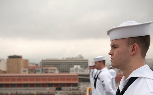 A group of U.S. Navy sailors in dress white uniforms are standing at attention on the deck of a naval vessel, likely during a formal manning-the-rails ceremony. The foreground sailor displays a rating badge on his left sleeve and service ribbons on his chest. The scene appears to be in an urban port environment under overcast skies.