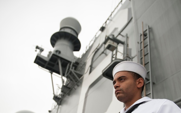 A U.S. Navy sailor in dress white uniform stands at attention on the deck of a naval warship, with a U.S. Marine in tan service uniform visible in the background. The sailor displays a Petty Officer rating badge on his left sleeve and a service ribbon on his chest. The image appears to depict a formal naval ceremony or manning the rails evolution.