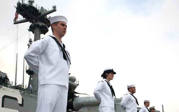 A line of U.S. Navy sailors in dress white uniforms are standing at attention along the rail of a naval vessel, performing a 'manning the rails' ceremonial formation. At least one individual in a Marine dress uniform (khaki/green) is also visible among the line. The ship's hull shows the number '6' and text reading 'AND ROTORS', suggesting this is a large amphibious or multi-purpose naval vessel.