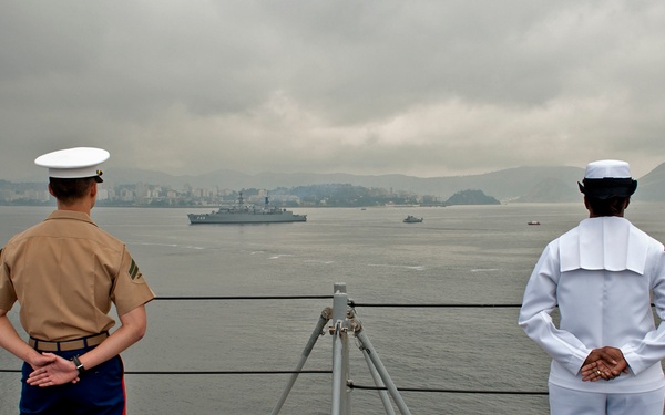 Two U.S. military personnel — a Marine in tan service uniform with blue trousers and a Navy sailor in dress whites — stand at parade rest on the deck of a naval vessel, observing a harbor with warships and a coastal city in the background. Both figures face away from the camera toward the water. The overcast sky and distant mountainous coastline suggest a port visit, possibly in Asia.