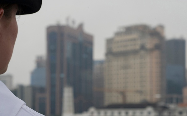 A female naval personnel member in white dress uniform is viewed from behind, looking out toward a dense urban waterfront skyline. A black shoulder patch on her uniform reads 'USS AMERICA', identifying her ship assignment. The cityscape in the background features multiple high-rise buildings along a harbor or port area.