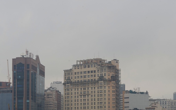 Military personnel in white dress uniforms and khaki/tan uniforms stand at parade rest along the edge of what appears to be a naval vessel's flight deck or upper deck, facing a city skyline. The formation suggests a formal 'manning the rails' ceremony, a traditional naval practice performed when entering or departing port. The urban backdrop features a mix of modern glass towers and older mid-century buildings consistent with a South American port city.