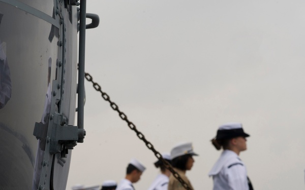 The image shows a group of U.S. Navy sailors and at least one U.S. Marine standing at attention on the deck of a naval vessel. The personnel are dressed in white service dress uniforms (sailors) and tan/khaki service uniform with red-striped trousers (Marine). A large anchor chain and gray ship hull components are visible in the foreground, with a hazy waterway and bridge structure visible in the background.