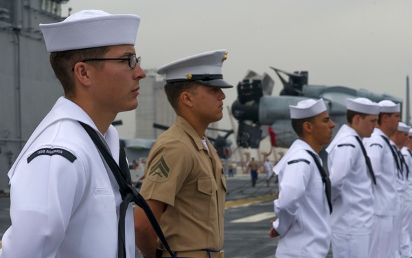 Military personnel are standing at attention in formation on the flight deck of a naval vessel, identified as the USS America based on the name tape visible on the foremost sailor's uniform. The formation includes U.S. Navy sailors in white dress uniforms and at least one U.S. Marine in service khaki uniform with blue dress trousers featuring a red stripe. Aircraft and ship superstructure are visible in the background, suggesting a formal ceremony or port arrival/departure event.