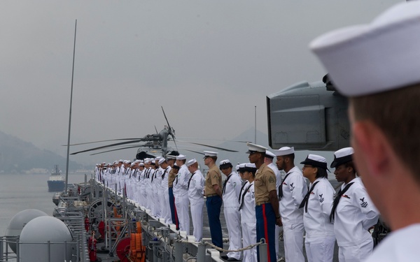 A formation of U.S. Navy sailors and Marines in dress uniforms are standing at attention along the rail of a naval warship, performing a 'manning the rails' ceremonial formation. The ship appears to be underway or approaching a port, with a mountainous coastline visible in the background through overcast skies. A helicopter with folded rotor blades is visible on the ship's deck.