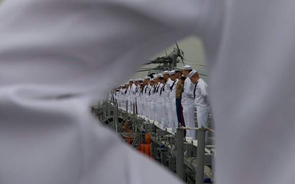 The image shows naval personnel in white dress uniforms standing in formation on the deck of a warship, viewed through the gap created by the arms of a foreground figure standing at parade rest. The creative framing uses the foreground sailor's white uniform sleeves and clasped hands to form a triangular window into the scene. At least one individual in a darker uniform (possibly a different branch or rank) is visible among the white-uniformed sailors.