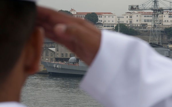 A naval personnel member in a white uniform is saluting toward a warship docked at a naval facility. The ship bears the hull number F41, identifying it as a frigate moored at what appears to be a naval base or shipyard. Industrial cranes, warehouses, and multi-story administrative buildings are visible in the background.