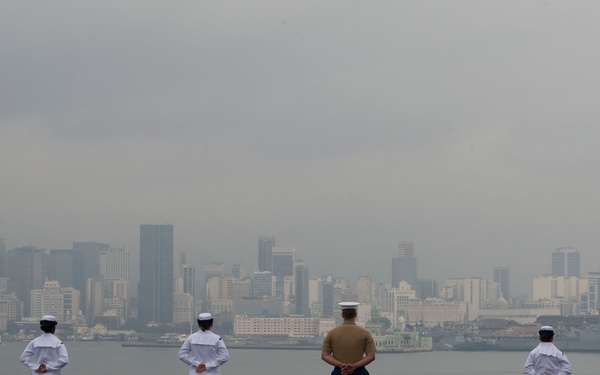 Military personnel are standing at parade rest on the flight deck of a naval vessel, facing a city skyline across a body of water. Six individuals in white Navy dress uniforms and one in a tan/khaki Marine Corps uniform are evenly spaced along the deck edge. The overcast sky and hazy atmosphere partially obscure the urban skyline in the background.