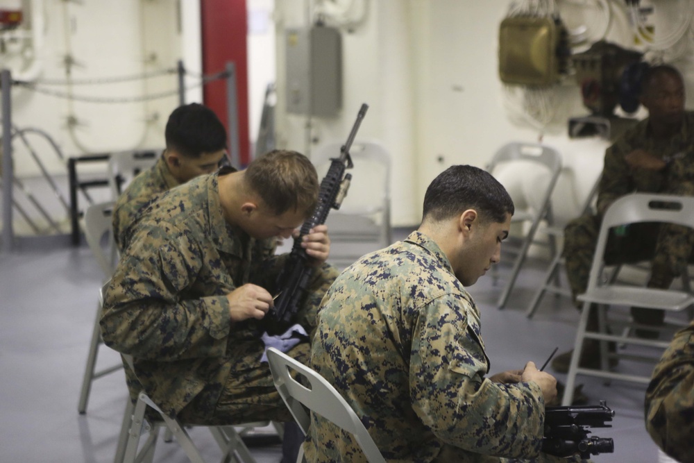 Several military personnel in MARPAT (Marine pattern) camouflage uniforms are seated on folding chairs in what appears to be an interior space of a naval vessel or military facility. The soldiers are engaged in weapons maintenance, cleaning and inspecting their rifles. The setting suggests a routine field maintenance session during a deployment or training exercise.
