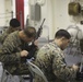 Several military personnel in MARPAT (Marine pattern) camouflage uniforms are seated on folding chairs in what appears to be an interior space of a naval vessel or military facility. The soldiers are engaged in weapons maintenance, cleaning and inspecting their rifles. The setting suggests a routine field maintenance session during a deployment or training exercise.