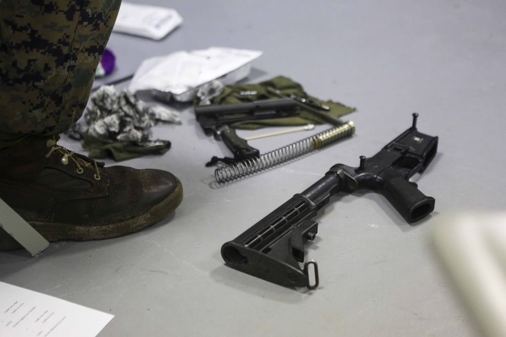 The image shows a disassembled rifle, consistent with an M16/AR-15 platform, laid out on a flat surface during what appears to be a field stripping or maintenance exercise. A person wearing MARPAT (Marine pattern) camouflage uniform and military combat boots is standing nearby. Cleaning supplies, tools, and components are spread across the surface.
