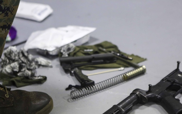 The image shows a disassembled rifle, consistent with an M16/AR-15 platform, laid out on a flat surface during what appears to be a field stripping or maintenance exercise. A person wearing MARPAT (Marine pattern) camouflage uniform and military combat boots is standing nearby. Cleaning supplies, tools, and components are spread across the surface.