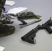 The image shows a disassembled rifle, consistent with an M16/AR-15 platform, laid out on a flat surface during what appears to be a field stripping or maintenance exercise. A person wearing MARPAT (Marine pattern) camouflage uniform and military combat boots is standing nearby. Cleaning supplies, tools, and components are spread across the surface.