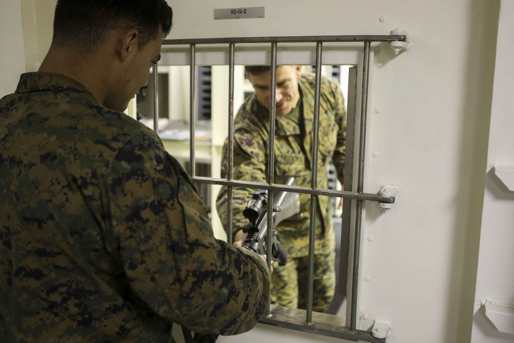 Two individuals in military camouflage uniforms are interacting through a barred window or pass-through opening in a white wall. One person on the near side is holding what appears to be a rifle or long firearm with a scope, passing or receiving it through the bars. A label reading '02-G-2' is visible above the barred opening.
