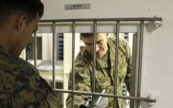 Two individuals in military camouflage uniforms are interacting through a barred window or pass-through opening in a white wall. One person on the near side is holding what appears to be a rifle or long firearm with a scope, passing or receiving it through the bars. A label reading '02-G-2' is visible above the barred opening.