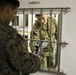 Two individuals in military camouflage uniforms are interacting through a barred window or pass-through opening in a white wall. One person on the near side is holding what appears to be a rifle or long firearm with a scope, passing or receiving it through the bars. A label reading '02-G-2' is visible above the barred opening.