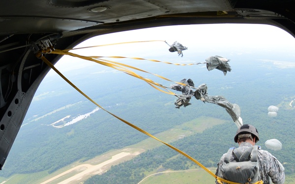 A military static-line parachute jump is in progress from the rear ramp of a large cargo aircraft, likely a CH-47 Chinook or C-130. A soldier in camouflage uniform is captured mid-exit, with yellow static lines deployed and parachutes opening in the background. The ground below shows a forested area with sandy drop zones and open fields consistent with a military training installation.