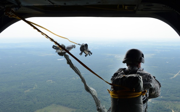 A military paratrooper is captured mid-exit from the rear ramp of a military aircraft, with their static line visibly deployed and parachute beginning to extract. The soldier is wearing full military combat uniform, helmet, and a packed parachute rig on their back. Below, a vast forested landscape with open fields and roads is visible from significant altitude.