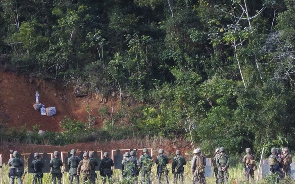 A group of military or security personnel in camouflage uniforms and tactical gear are lined up at an outdoor shooting range. The range features multiple dark silhouette targets mounted on a frame, set against a red-earth embankment serving as a backstop. The setting appears to be a jungle or tropical forest environment.