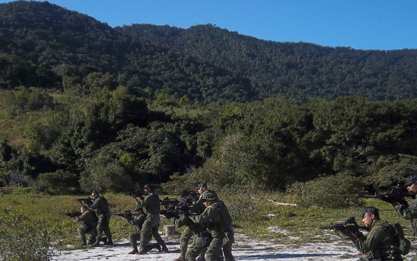 A group of military or law enforcement personnel in green camouflage uniforms are conducting a tactical field training exercise on open sandy terrain. The personnel are positioned in a line formation, aiming rifles in a coordinated drill. The setting is a rural outdoor area with dense forested hills in the background under clear blue skies.