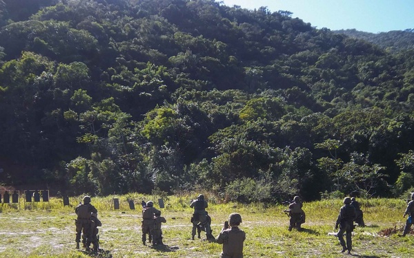 The image shows a group of military or law enforcement personnel conducting a live-fire or tactical training exercise on an outdoor shooting range. Multiple individuals in camouflage uniforms and helmets are positioned across a grassy field, facing a row of silhouette targets set against a densely forested hillside. The setting appears to be a tropical or subtropical environment based on the vegetation.
