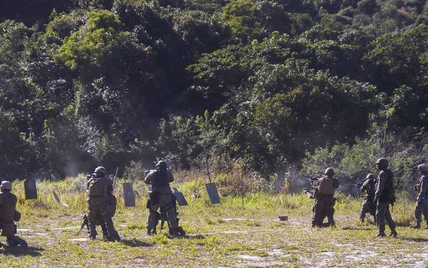 A group of military or law enforcement personnel in full tactical gear are conducting a live-fire training exercise on an outdoor range. Multiple shooters are engaged simultaneously against dark metal silhouette targets, with visible smoke/muzzle discharge evident at several positions. The setting is a cleared grassy field bordered by dense tropical or subtropical vegetation.