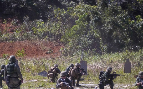 A group of approximately 8-10 armed military personnel are conducting what appears to be a live-fire training exercise on an outdoor range. Soldiers are in various tactical positions including kneeling, crouching, and standing, with weapons raised toward silhouette targets downrange. The setting is a cleared field area bordered by dense tropical or subtropical vegetation and red clay embankments.