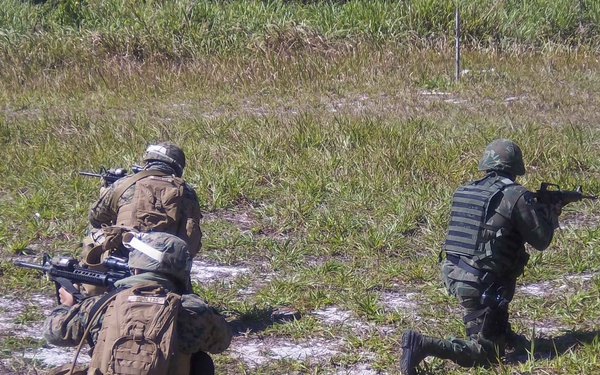 The image shows three armed military personnel conducting what appears to be a tactical field exercise in an open grassy area. Two soldiers on the left wear digital camouflage (MARPAT-style) uniforms with tactical gear and backpacks, while a third on the right wears woodland camouflage. All three are in low crouching or kneeling positions and are carrying rifles, suggesting a coordinated movement or assault drill.
