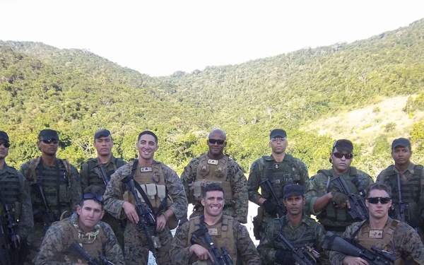A group of approximately 13 armed military personnel pose for a group photograph in an outdoor setting with forested hills in the background. The group appears to consist of members from at least two different military forces, identifiable by their distinct camouflage uniform patterns. They are equipped with rifles and tactical gear, suggesting this may be a joint military training exercise or operation.