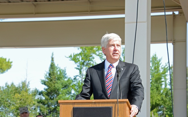 A silver-haired man in a dark suit with a red, white, and blue striped tie is delivering a speech at an outdoor podium on a wooden stage. The setting appears to be a patriotic or military ceremony, evidenced by the American bunting draped across the front of the stage and uniformed military personnel visible in the background audience.