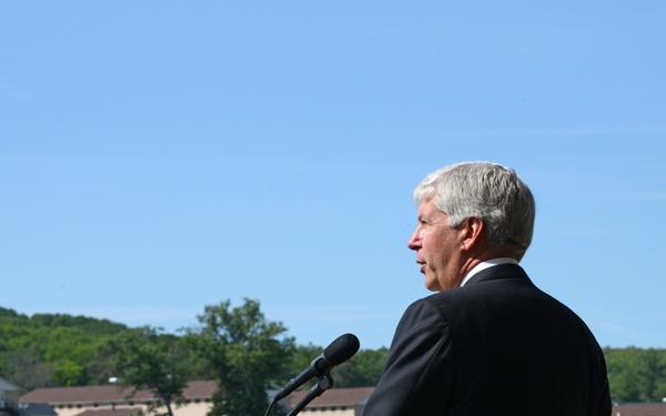 A civilian man in a dark suit is speaking at a wooden podium with a microphone during an outdoor military ceremony. A large formation of uniformed soldiers stands at attention on a parade field in the background. The setting appears to be a military installation with barracks-style buildings and wooded hills visible behind the troops.