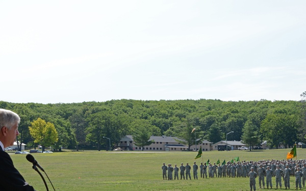 A civilian official in a dark suit is speaking at a wooden podium during a military formation ceremony on a large grass parade field. A large formation of uniformed military personnel stands at attention in the background, with unit guidon flags visible among the ranks. The setting appears to be a military installation with buildings and tree lines visible in the distance.