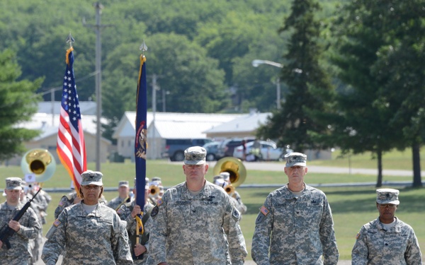 A formation of U.S. Army soldiers in ACU (Army Combat Uniform) pattern camouflage marches down a gravel road at what appears to be a military installation. A color guard carries the U.S. flag and what appears to be a unit or state flag, while a military brass band follows behind. The setting is a green, tree-lined base with buildings visible in the background.