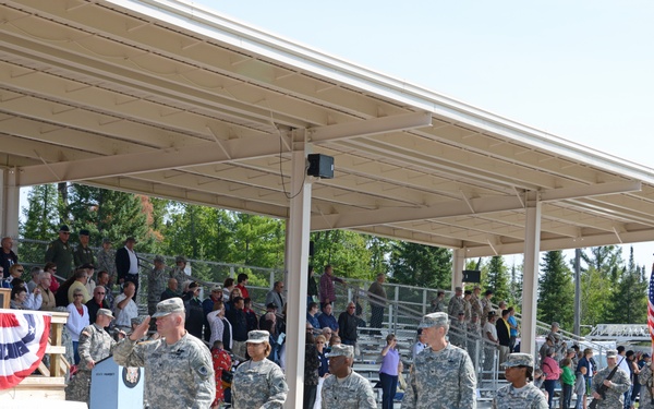 A military ceremony is taking place at an outdoor covered pavilion or grandstand area. Soldiers in ACU (Army Combat Uniform) camouflage are marching or parading past a reviewing stand decorated with patriotic red, white, and blue bunting. A crowd of civilians and military personnel observes from bleachers under the covered structure.
