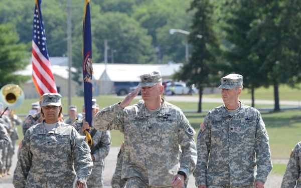 A group of U.S. Army soldiers in ACU (Army Combat Uniform) digital camouflage are marching in formation along a gravel path at what appears to be a military installation. A central figure is rendering a salute while leading the formation, accompanied by other soldiers including flag bearers carrying the American flag and a unit guidon. A military band with brass instruments is visible in the background.