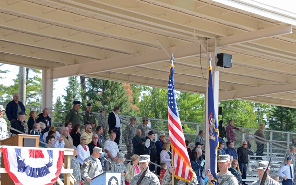 A military color guard ceremony is taking place at an outdoor covered pavilion or amphitheater. Soldiers in Army Combat Uniforms (ACU) are marching with the U.S. flag and what appears to be a unit or state flag, while a crowd of civilians and military personnel observe from bleachers. A podium with patriotic bunting decorations is visible on a raised platform, suggesting a formal military or veterans ceremony.