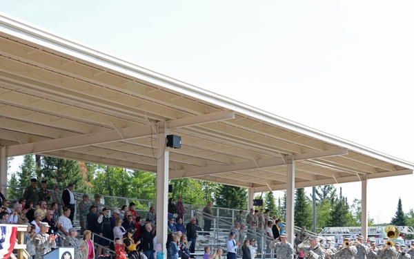 A military ceremony is taking place at an outdoor covered pavilion or grandstand, with a formation of uniformed soldiers and a military band performing. A podium with patriotic red, white, and blue bunting is visible on a raised platform to the left, suggesting an official address or ceremony. Civilians and military personnel fill the bleachers as spectators.