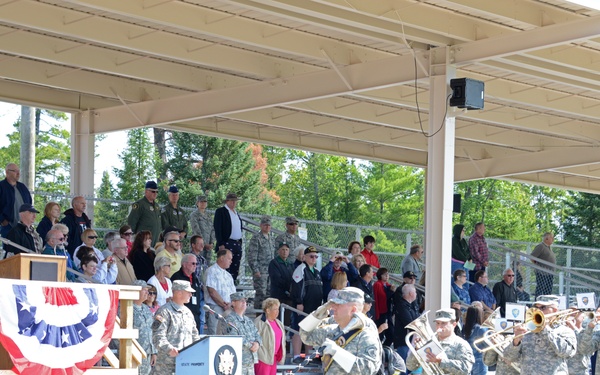 A military ceremony is taking place at an outdoor covered pavilion, featuring a uniformed military band marching and playing brass instruments. A podium with patriotic red, white, and blue bunting decorations is visible on a raised stage, with dignitaries and civilians watching from bleachers. The event appears to be a formal military commemoration or dedication ceremony.