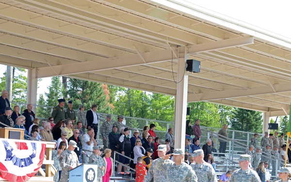 A military ceremony is taking place at an outdoor covered pavilion with bleacher seating. Soldiers in Army Combat Uniforms (ACU) are marching in formation past a speaking platform decorated with patriotic bunting, while civilians and military personnel observe from the stands. A green unit flag/guidon is visible being carried during what appears to be a formal military event such as a deployment ceremony, homecoming, or change of command.