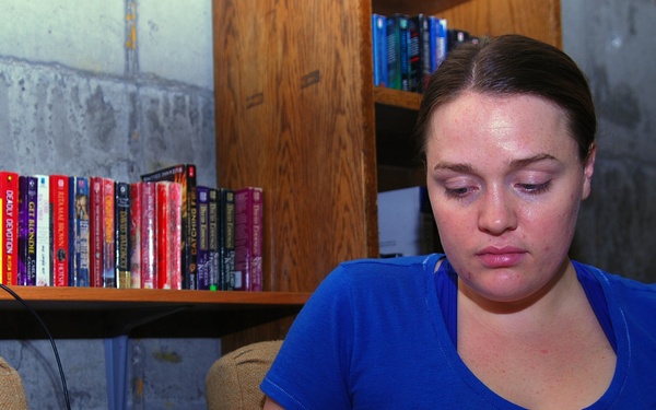 A young woman with dark hair pulled back is seated in a beige armchair reading a paperback book. She is wearing a blue t-shirt and appears focused on her reading. The setting appears to be a basement or utility room with concrete walls, a wooden bookshelf filled with numerous paperback books visible in the background.