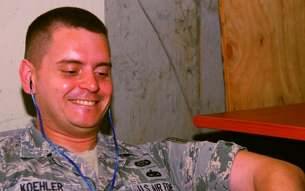 A U.S. Air Force servicemember named Koehler, wearing Airman Battle Uniform (ABU), is seated in a chair smiling while using a tablet device. He has blue earbuds/earphones in his ears, suggesting he is watching or listening to content. The setting appears to be an informal relaxation or common area, possibly at a deployed location.