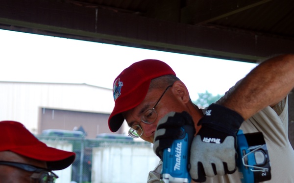 Two workers wearing matching red caps and work gloves are collaborating on a construction or carpentry task under a covered outdoor structure. One man is operating a blue Makita cordless drill into a plywood surface while the other observes closely from a lower angle. Both are wearing safety glasses and appear to be engaged in focused, cooperative work.