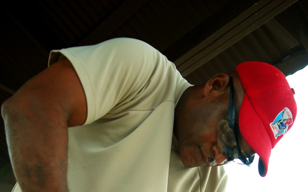 A man wearing US Air Force digital camouflage (ABU) pants and a red baseball cap is using a blue Makita cordless drill on a wooden surface. He is leaning forward intently while wearing black work gloves, suggesting he is engaged in a construction or repair task. The setting appears to be under a covered outdoor structure.